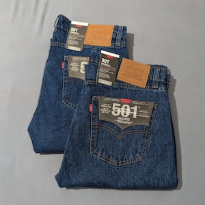 May include: Two pairs of dark blue denim jeans with the "501 Original" label. Each pair has a brown leather patch and a tag with product information. The jeans are folded and laid flat on a gray surface.