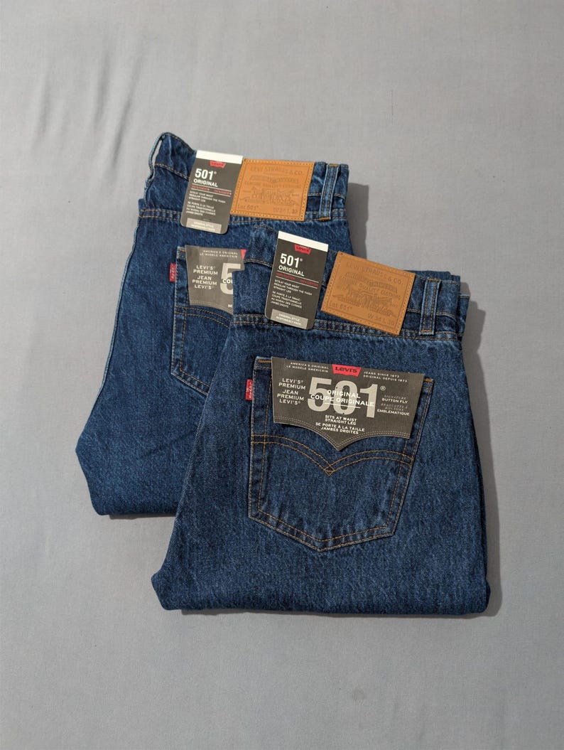 May include: Two pairs of dark blue denim jeans with the "501 Original" label. Each pair has a brown leather patch and a tag with product information. The jeans are folded and laid flat on a gray surface.