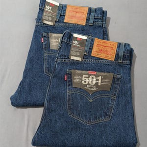 May include: Two pairs of dark blue denim jeans with tags. The jeans have the "501 ORIGINAL" label and a red Levi's tag on the back pocket. The jeans are folded neatly, showcasing the back pockets and the brand labels.