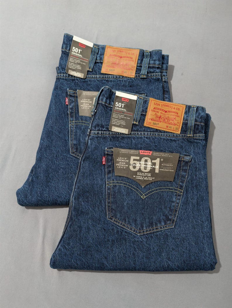 May include: Two pairs of dark blue denim jeans with tags. The jeans have the "501 ORIGINAL" label and a red Levi's tag on the back pocket. The jeans are folded neatly, showcasing the back pockets and the brand labels.