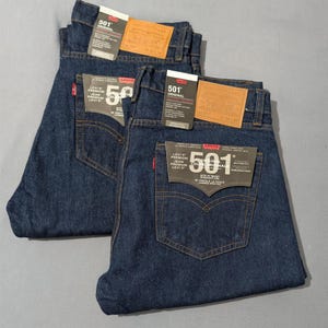 May include: Two pairs of dark blue Levi's 501 Original jeans, folded neatly. Each pair has a brown leather patch and a black tag with white text. The jeans are made of denim and have classic five-pocket styling.