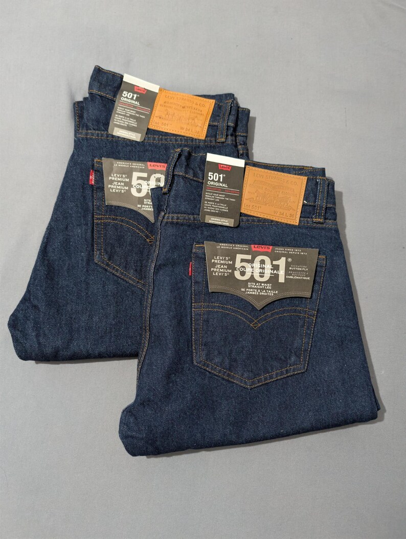May include: Two pairs of dark blue Levi's 501 Original jeans, folded neatly. Each pair has a brown leather patch and a black tag with white text. The jeans are made of denim and have classic five-pocket styling.