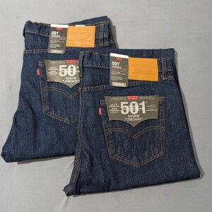May include: Two pairs of dark wash denim jeans with the Levi's 501 label visible. The jeans have classic five-pocket styling, copper rivets, and a red Levi's tag. The jeans are folded and laid flat on a gray surface.