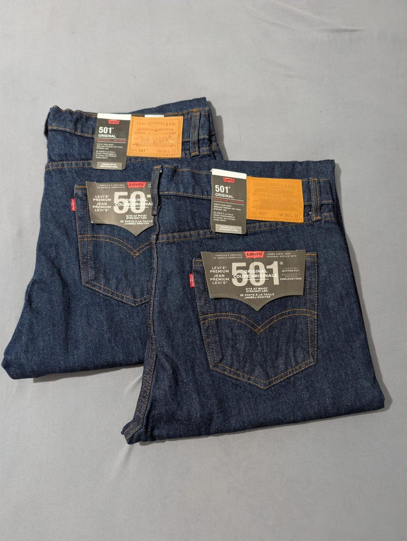 May include: Two pairs of dark wash denim jeans with the Levi's 501 label visible. The jeans have classic five-pocket styling, copper rivets, and a red Levi's tag. The jeans are folded and laid flat on a gray surface.