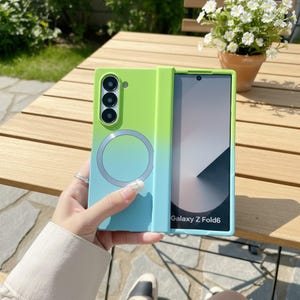 Könnte beinhalten: Eine grün-blaue Handyhülle mit Farbverlauf für ein Galaxy Z Fold6. Die Hülle hat einen kreisförmigen Magnetring und einen Ausschnitt für eine Dreifachkamera. Der Telefonbildschirm zeigt eine schwarz-weiße Grafik.