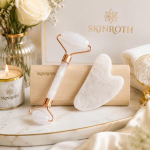 Puede incluir: Un rodillo facial de cuarzo blanco con herrajes de metal en oro rosa y una piedra gua sha a juego en forma de corazón. Ambos artículos tienen la marca SKINROTH grabada en el mango y la superficie de la piedra, sobre un bloque de madera clara.