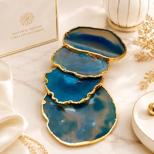 Könnte beinhalten: Ein Set aus vier unregelmäßigen, kreisförmigen Achat-Untersetzern mit tiefblauen und durchscheinenden weißen Bändern sowie goldfarben lackierten Rändern. Die Untersetzer sind vertikal gestapelt und zeigen natürliche Steinmuster und polierte Oberflächen.