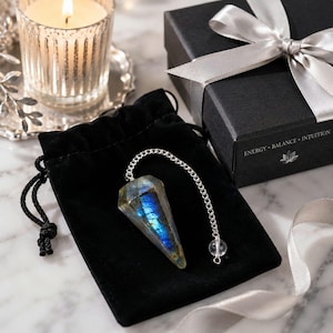 May include: A faceted labradorite pendulum with a metallic chain and a small clear bead at the end, resting on a black velvet drawstring pouch. Beside it sits a black gift box with a silver ribbon and the text ENERGY BALANCE INTUITION.