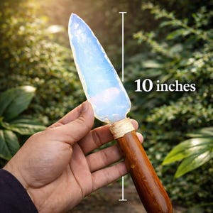 Puede incluir: Un cuchillo artesanal de opalita con mango de madera. La hoja es translúcida, azul pálido, y el mango está envuelto con una fibra natural. El cuchillo mide 25,4 cm de largo. El cuchillo se sostiene contra un fondo de follaje verde.