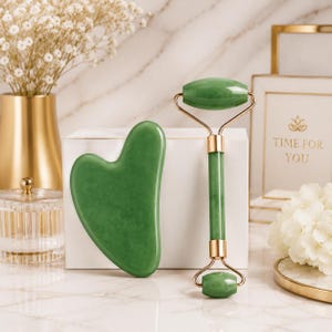 Puede incluir: Un rodillo facial de jade verde y una herramienta gua sha en forma de corazón. El rodillo presenta una piedra cilíndrica grande en la parte superior y una más pequeña en la inferior, unidas por herrajes de metal dorado. Ambos tienen acabado liso.
