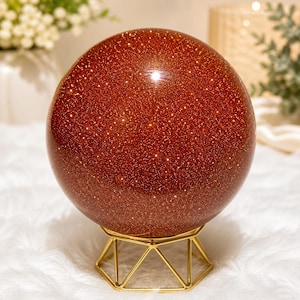 May include: A large, sparkling, reddish-brown sphere, possibly a decorative crystal ball, sits on a gold-colored geometric stand. The sphere has a glittery appearance, with tiny reflective particles throughout. The stand has a hexagonal base.