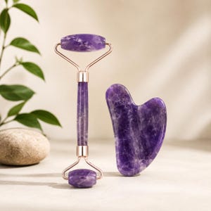 Puede incluir: Un rodillo facial de doble extremo y una herramienta gua sha en forma de corazón, ambos hechos de piedra amatista de color púrpura intenso con vetas blancas naturales. El rodillo cuenta con herrajes de metal en tono oro rosa.