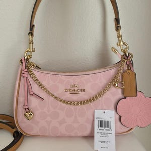 Könnte beinhalten: Eine hellrosa Coach-Schultertasche mit einem charakteristischen Monogramm-Muster, goldfarbenen Beschlägen, einem abnehmbaren Goldkettengurt und einem beigen Lederanhänger. Ein rosafarbener blütenförmiger Anhänger und ein kleines Herz sind befestigt.