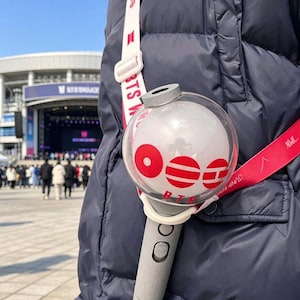 Puede incluir: Un bastón de luz BTS gris y blanco con una parte superior esférica transparente con gráficos rojos circulares y de líneas horizontales, el texto BTS y una correa blanca con la marca BTS en rojo, sobre una chaqueta de invierno acolchada azul marino.