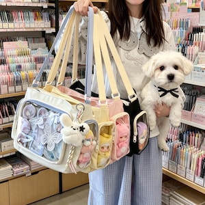 May include: A collection of three multi-compartment tote bags in white, yellow, and black, each featuring a clear front window display pocket, pastel-coloured straps, and a fluffy white rabbit-shaped keychain attached to the side.