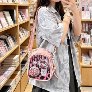 Könnte beinhalten: Eine hellrosa Umhängetasche mit einer durchsichtigen Vordertasche, in der zwei runde Anstecknadeln im Anime-Stil und eine kleine rechteckige Karte zu sehen sind. Die Tasche hat einen passenden verstellbaren Schultergurt und ein schwarz-rosa Kuhmuster-Innenfutter.