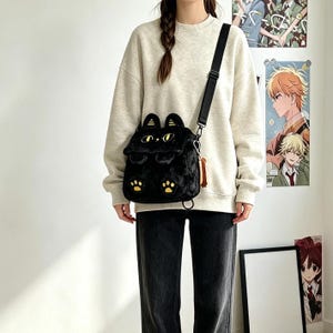 May include: A black plush cat-shaped crossbody bag featuring yellow eyes, small ears, and yellow paw prints on the front. The bag has a black adjustable shoulder strap and is worn over a beige crewneck sweatshirt paired with dark gray denim jeans.