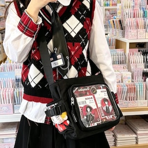 Könnte beinhalten: Eine schwarze Umhängetasche mit einer durchsichtigen Vordertasche, die rot-weiße Grafikkarten mit Anime-Charakterillustrationen zeigt. Die Tasche verfügt über eine Seitentasche mit einer kleinen Plüschfigur und einen verstellbaren Riemen.