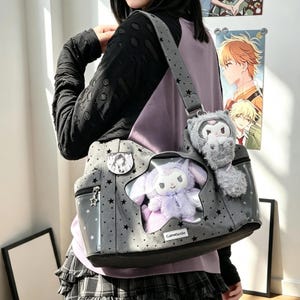 May include: A grey shoulder bag featuring a star-patterned fabric, a star-shaped clear window displaying a plush purple character, and a grey plush charm attached to the strap. The bag includes a small rectangular label with the text GameSouls.