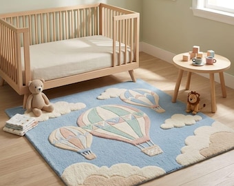 May include: A rectangular area rug featuring a light blue background with a tufted design of three hot air balloons in shades of sage green, dusty pink, and cream, surrounded by fluffy white clouds, placed on a light wood floor in a room.