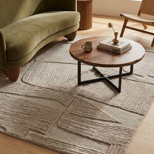 May include: A living room scene featuring a curved olive green velvet sofa, a round wooden coffee table with a black metal base, and a textured beige rug. A small side table and a woven chair are also visible.