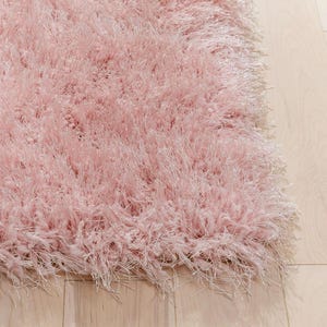 Peut inclure: Gros plan sur un tapis rose doux et duveteux. Le tapis a de longues fibres hirsutes, cr&eacute;ant une texture moelleuse. La couleur est un rose clair pastel, et le tapis semble neuf et propre. Le tapis est sur un plancher de bois clair.