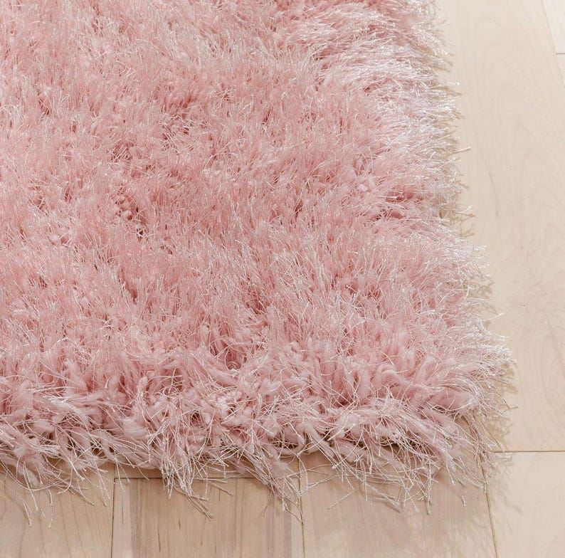 Peut inclure: Gros plan sur un tapis rose doux et duveteux. Le tapis a de longues fibres hirsutes, cr&eacute;ant une texture moelleuse. La couleur est un rose clair pastel, et le tapis semble neuf et propre. Le tapis est sur un plancher de bois clair.