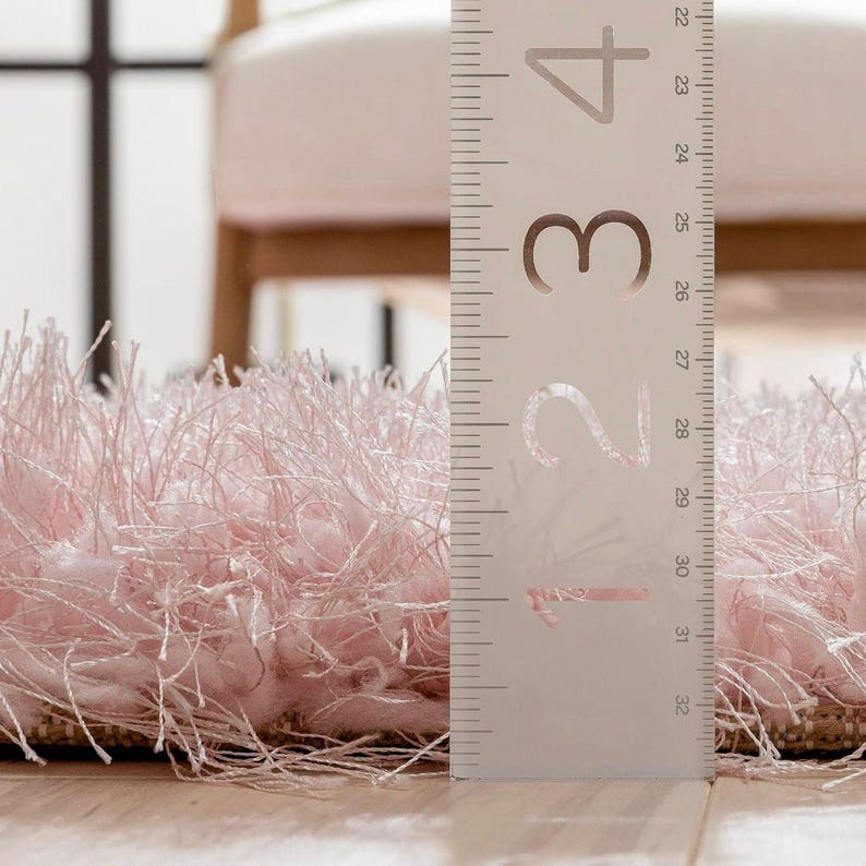 Peut inclure: Gros plan sur un tapis rose clair et duveteux avec une texture hirsute. Une r&egrave;gle est plac&eacute;e verticalement &agrave; c&ocirc;t&eacute; du tapis, indiquant des mesures de 56 &agrave; 81 cm. Les fibres du tapis sont longues et cr&eacute;ent un aspect doux et textur&eacute;.