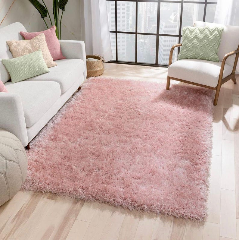 Peut inclure: Un grand tapis rose clair et duveteux recouvre le sol. La texture du tapis est hirsute, cr&eacute;ant une apparence douce et accueillante. Un canap&eacute; blanc avec des coussins color&eacute;s et un fauteuil blanc sont plac&eacute;s sur le plancher en bois clair, cr&eacute;ant un espace de vie confortable.