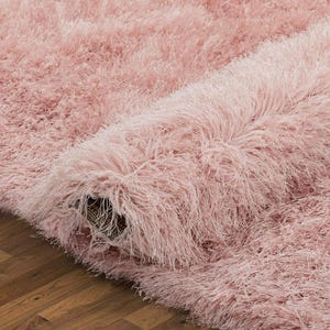 Peut inclure: Gros plan sur un tapis rose pelucheux enroul&eacute;. Le tapis a une texture hirsute avec de longues fibres douces. Le tapis est partiellement enroul&eacute;, r&eacute;v&eacute;lant le dessous et le sol en bois en dessous.