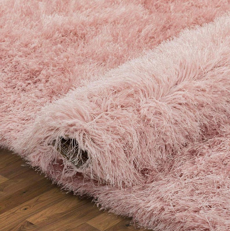 Peut inclure: Gros plan sur un tapis rose pelucheux enroul&eacute;. Le tapis a une texture hirsute avec de longues fibres douces. Le tapis est partiellement enroul&eacute;, r&eacute;v&eacute;lant le dessous et le sol en bois en dessous.