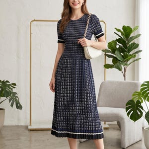 Può includere: Un abito midi blu navy con stampa a motivi bianchi, scollo rotondo, maniche corte con polsini a righe bianche e nere, corpetto arricciato e gonna plissettata con orlo a righe bianche e nere, abbinato a una borsa a spalla bianca.