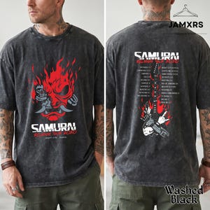 May include: A washed black t-shirt featuring a red and white graphic of a robotic samurai mask on the front with the text SAMURAI REUNION TOUR 2020. The back displays a guitar graphic with flames and a list of tour cities under the text SAMURAI REUNION TOUR 2020.