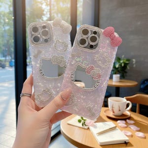 May include: Two clear phone cases with a Hello Kitty design. One case is light pink, the other is white. Both cases have a bow at the top and are decorated with rhinestones. The cases have a pearlescent finish and a cut-out of Hello Kitty's face.