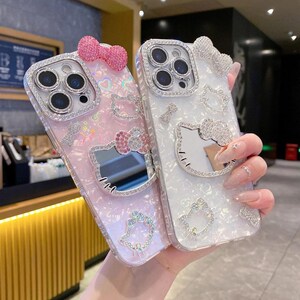 May include: Two decorative phone cases. One is pink with a bow, a Hello Kitty design, and rhinestones. The other is white with a bow, a Hello Kitty design, and rhinestones. Both cases have a mirrored surface.