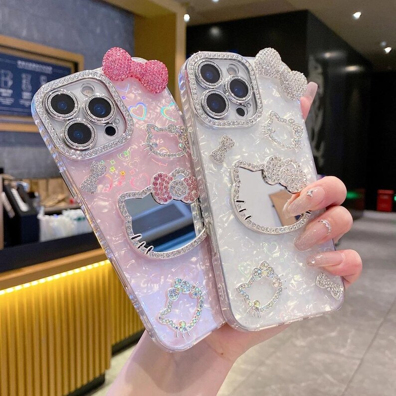 May include: Two decorative phone cases. One is pink with a bow, a Hello Kitty design, and rhinestones. The other is white with a bow, a Hello Kitty design, and rhinestones. Both cases have a mirrored surface.