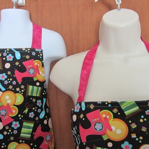 Könnte beinhalten: Zwei schwarze Schürzen mit rosa Trägern und einem bunten Nähmaschinen-Druck. Der Druck enthält Knöpfe, Garnrollen, Scheren und andere Nähutensilien.