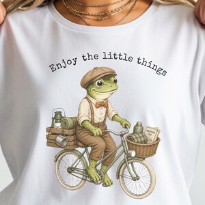 May include: White t-shirt with the text "Enjoy the little things." The shirt features a whimsical illustration of a frog in a cap and bow tie riding a bicycle with camping gear. The bicycle is green.
