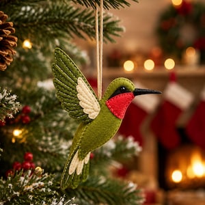 Puede incluir: Un adorno de fieltro con forma de colibrí que presenta alas y cuerpo de color verde oliva, una mancha roja brillante en la garganta y detalles en color crema en las alas y la cola. El ave tiene un pico negro fino y cuelga de un cordel marrón.