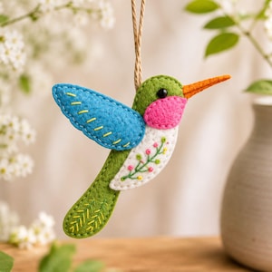 Puede incluir: Un adorno de fieltro con forma de colibrí que presenta un ala azul brillante con costuras amarillas, cabeza y cola verdes, una mancha rosa en la garganta y un vientre blanco con bordado floral. El ave tiene un pico naranja y cuelga de un cordel.