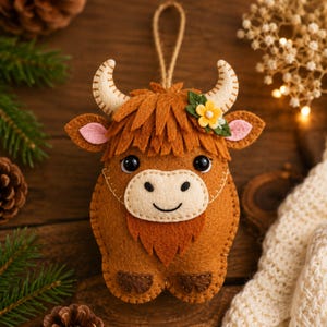 May include: A felt Highland cow ornament featuring a brown body, cream-colored muzzle, and curved horns. The cow has black bead eyes, a stitched smile, and a small yellow flower accent on its head. A jute twine loop is attached to the top for hanging.