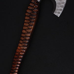以下が含まれることがあります： 銀色のテクスチャード加工されたヘッドと濃い茶色の木製ハンドルを備えた手作りの斧。ハンドルには詳細な彫刻模様があります。斧は黒い背景に配置されています。