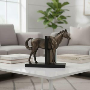 May include: Bronze-colored horse bookends. The horse statue stands between two black bookends, holding books in place. The horse is on a white table in front of a gray couch and a window.
