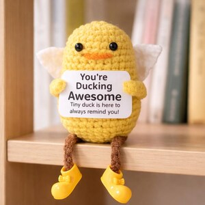 Op de afbeelding: Een gehaakte gele eend met zwarte ogen, een oranje snavel en kleine witte vleugels. De eend houdt een bord vast met de tekst "You're Ducking Awesome". Het heeft bruine poten en draagt gele schoenen met strikken.