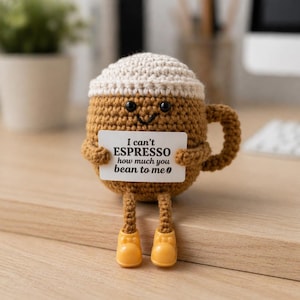 Puede incluir: Una taza de caf&eacute; tejida a ganchillo con una cara sonriente, sosteniendo un letrero que dice "I can't ESPRESSO how much you bean to me". La taza es marr&oacute;n con una parte superior color crema y zapatos amarillos. La taza tiene un asa y est&aacute; sobre una superficie de madera.