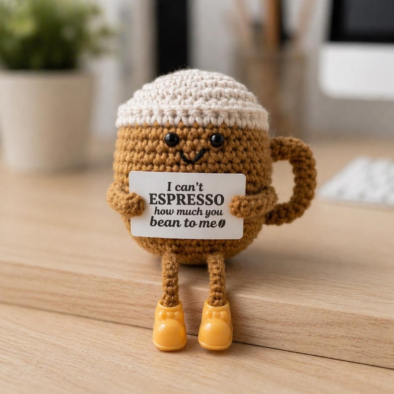 Puede incluir: Una taza de caf&eacute; tejida a ganchillo con una cara sonriente, sosteniendo un letrero que dice "I can't ESPRESSO how much you bean to me". La taza es marr&oacute;n con una parte superior color crema y zapatos amarillos. La taza tiene un asa y est&aacute; sobre una superficie de madera.