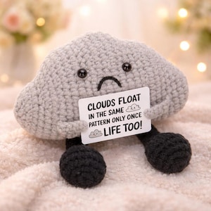 May include: A crocheted grey cloud-shaped plush toy with black button eyes and a stitched frown. It holds a white sign that reads "Clouds float in the same pattern only once, life too!" The toy has two black spherical feet.