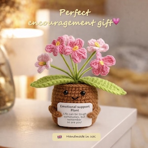 Könnte beinhalten: Eine handgefertigte Häkelpflanze mit rosa Blüten und grünen Blättern in einem braunen Topf. Der Topf hat ein lächelndes Gesicht und hält ein Schild mit der Aufschrift "Emotional support Plant". Der Text "Handmade in UK" ist ebenfalls sichtbar.