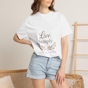 以下が含まれることがあります： 「Live Simply」の文字が筆記体で書かれた白いTシャツ。周りには花の模様があります。「Cherish the Little Things」というフレーズが下にあります。モデルはデニムのショートパンツを着用しています。