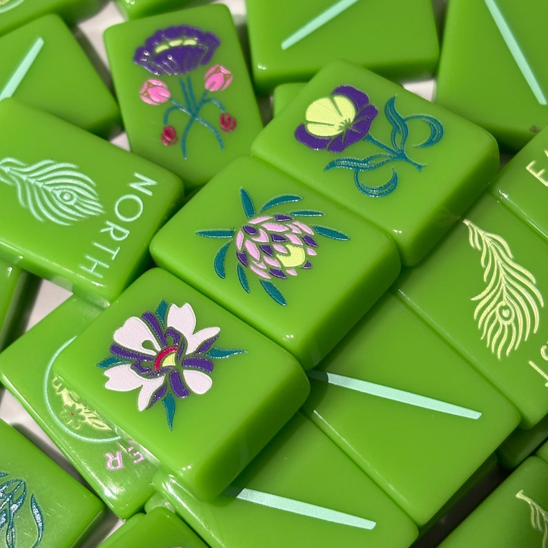 May include: A collection of bright green mahjong tiles featuring various engraved designs including colorful floral illustrations, a peacock feather, and the word NORTH in white lettering, all set against a smooth, glossy surface.